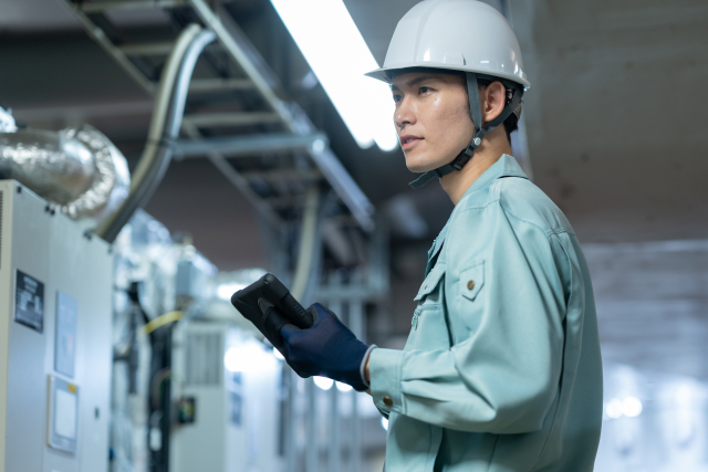 工場でタブレットを手に設備を見つめるヘルメット姿の作業者